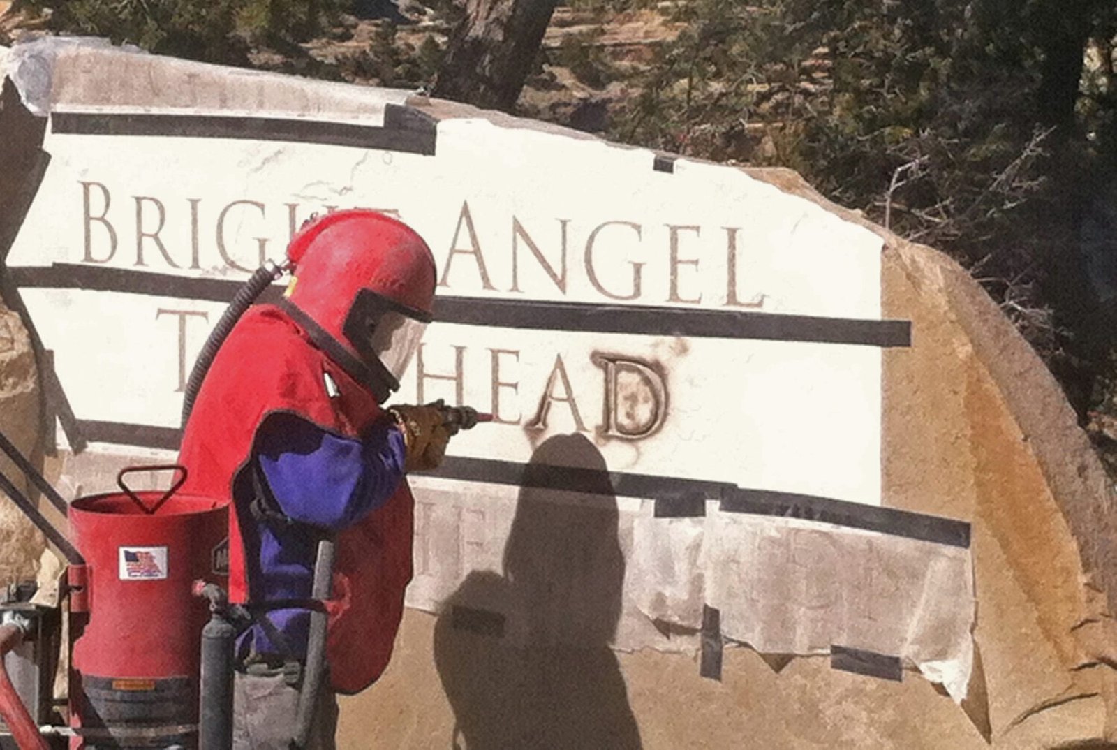 A worker in a red hood and blast suit sandblasting letters through a stencil